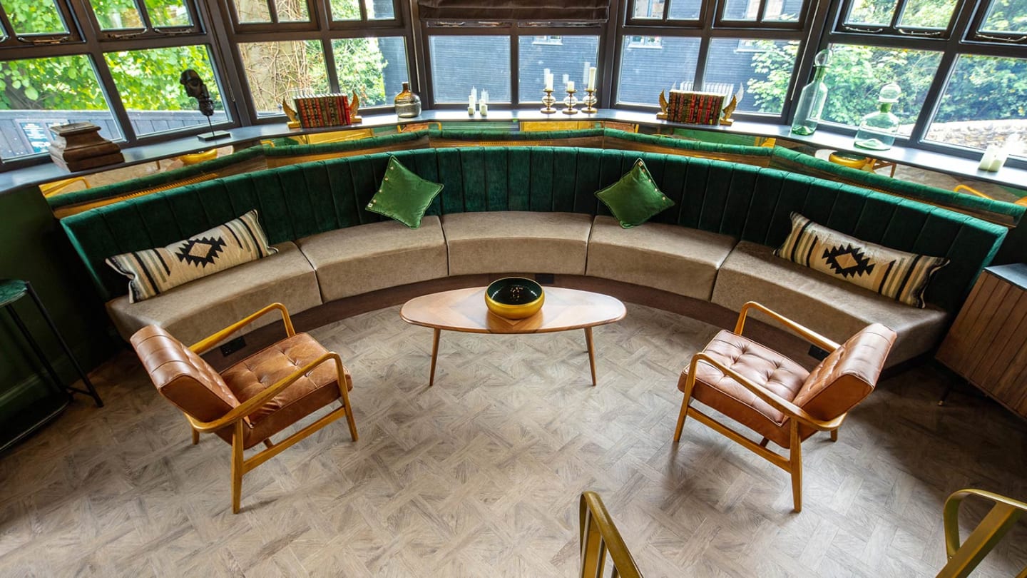 Circular seating area features warm oak inspired flooring with green velvet cushions and brown furniture.
