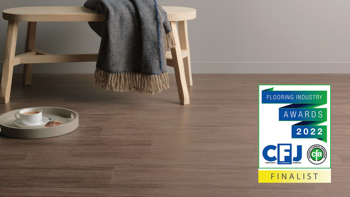 Brown wood-effect Amtico flooring with a light-coloured bench and grey blanket. Image includes a CFJ Flooring Industry Awards 2022 Finalist logo.